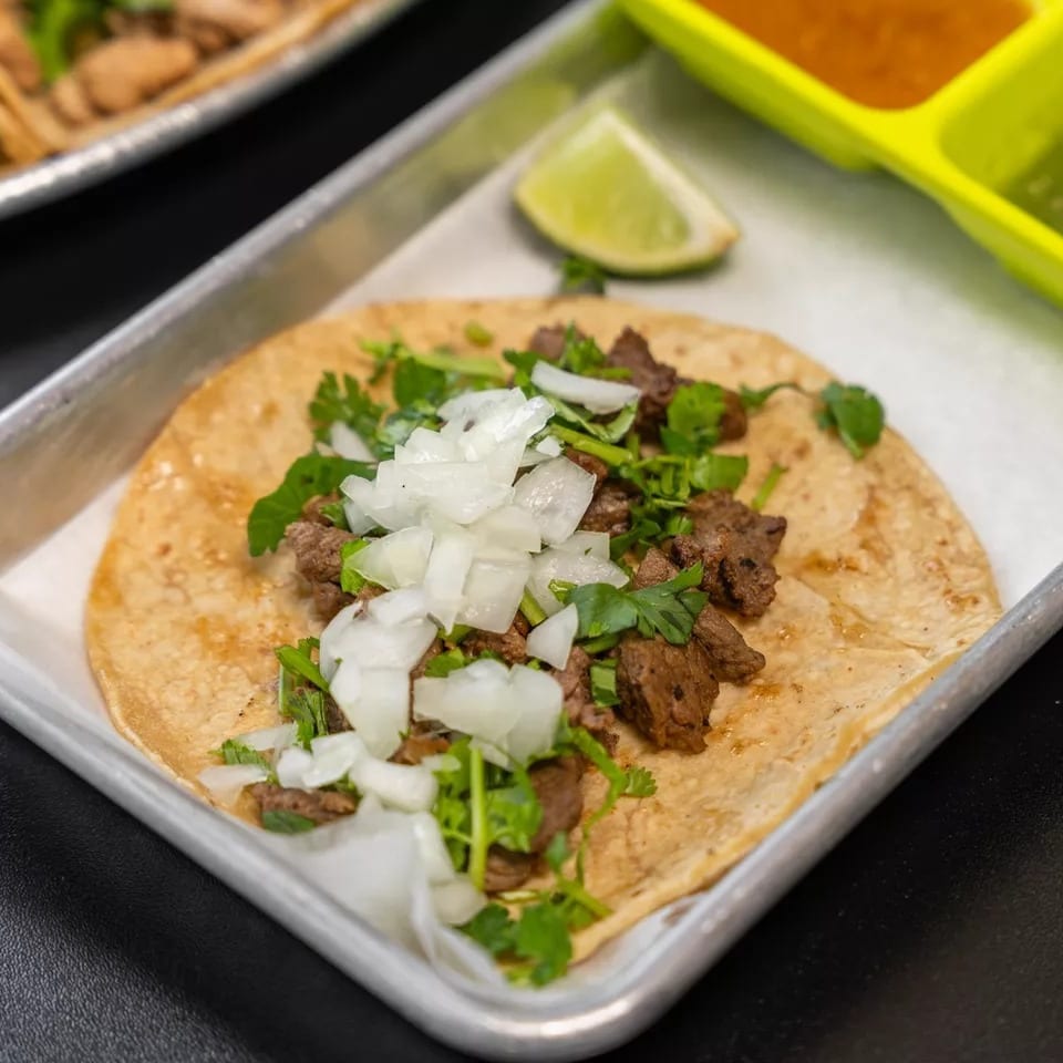 A single carne asada taco with onions and cilantro on a metal tray with lime and sauces.