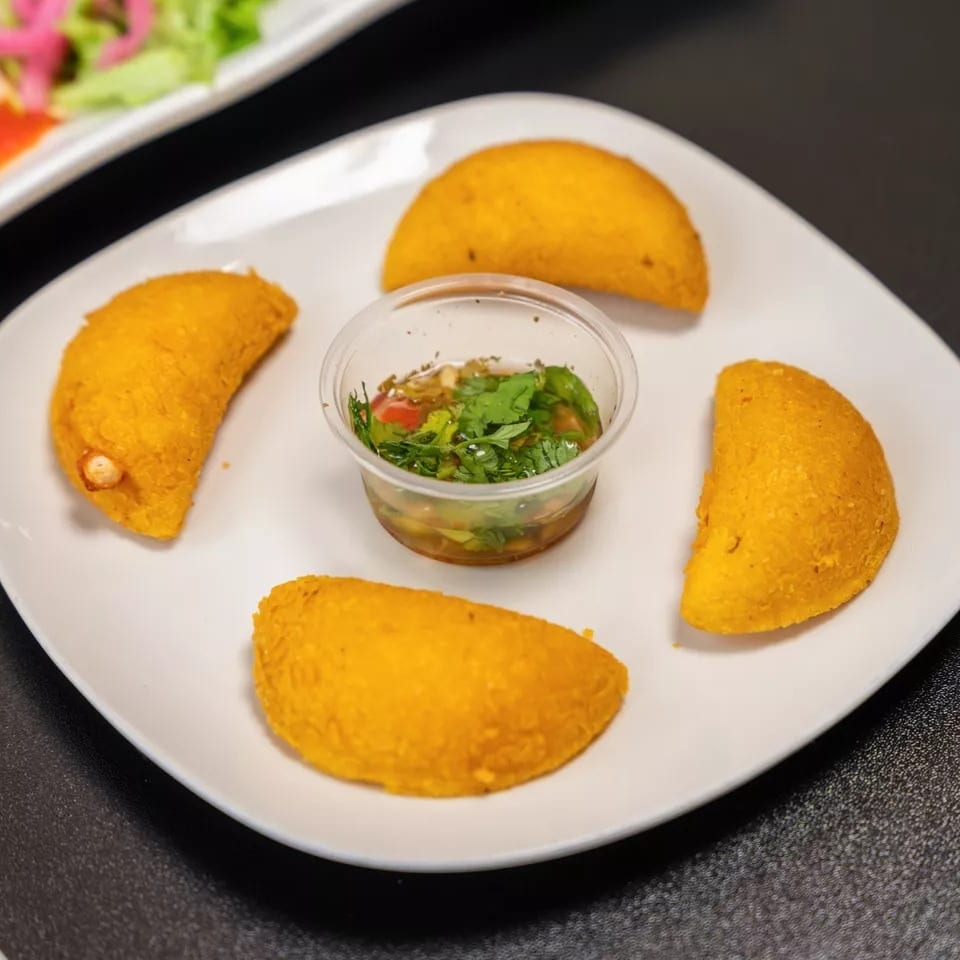 Four golden empanadas arranged around a small cup of green salsa on a white plate.