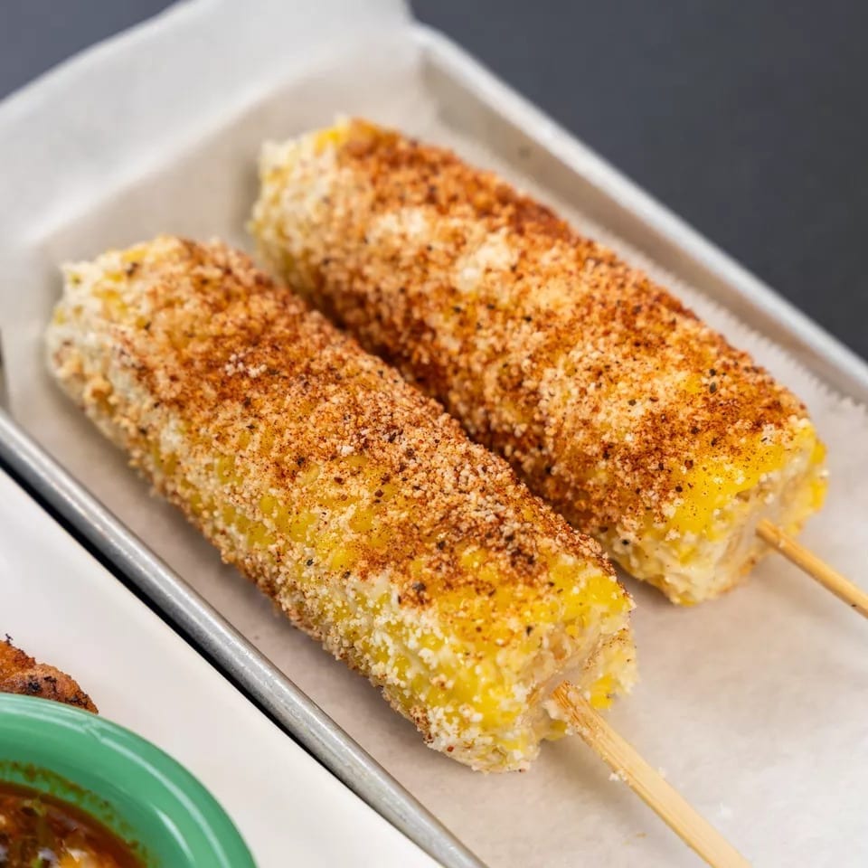 Two elotes, Mexican street corn on the cob, covered in cheese and chili powder on a tray.