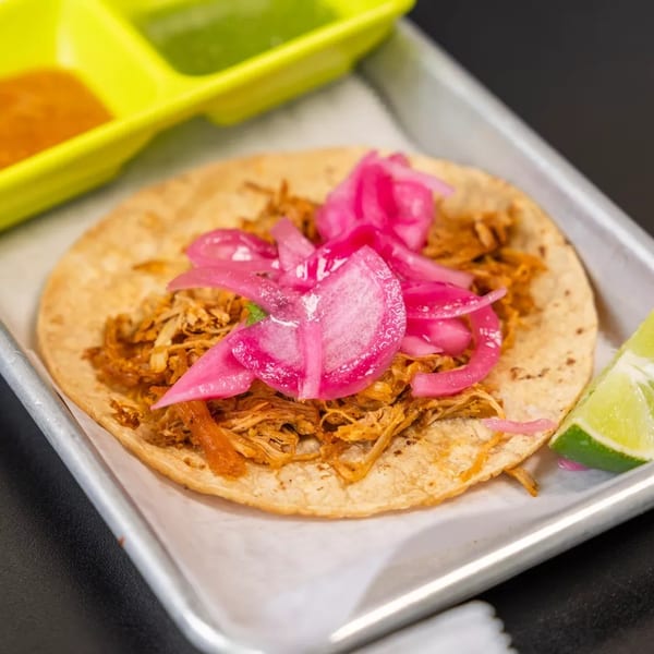 A single taco with shredded meat and pink pickled onions, served with lime and sauces.
