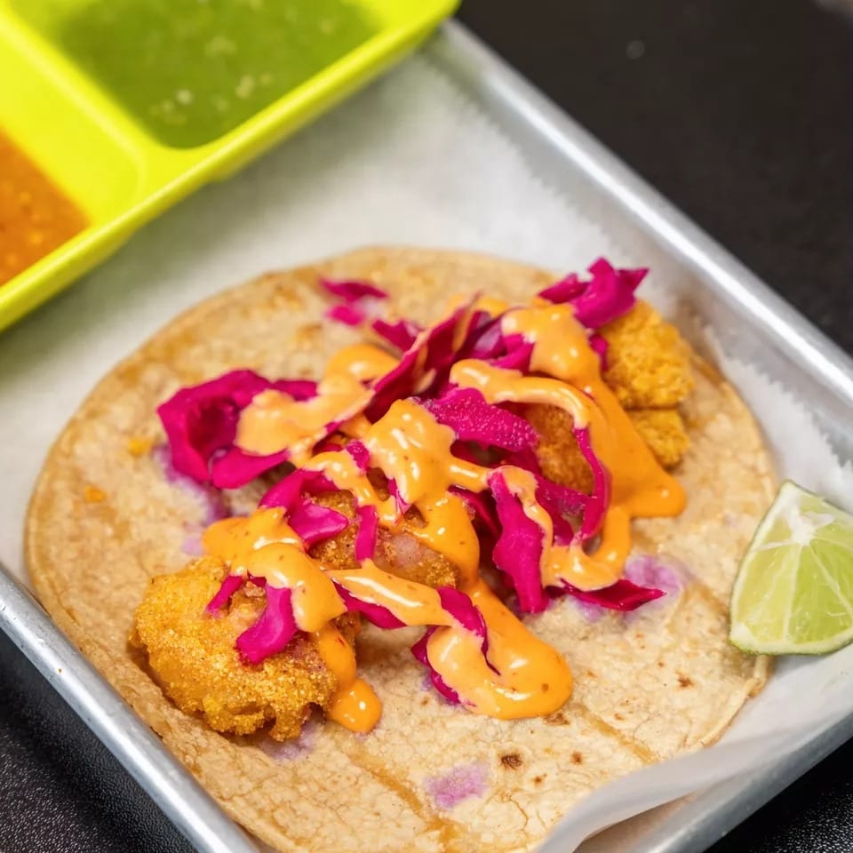 A shrimp taco with red cabbage and orange sauce, served with a lime wedge and two dipping sauces.
