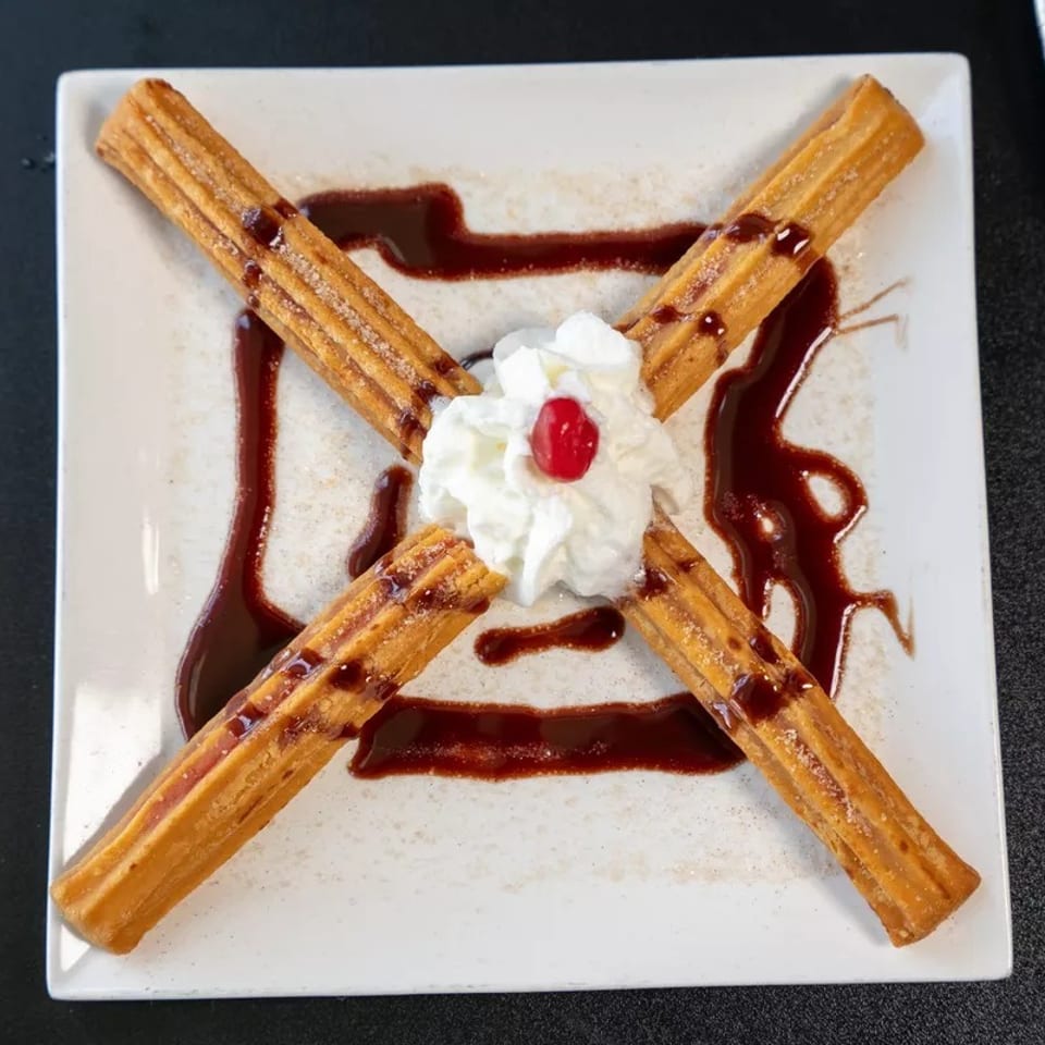 Four churros arranged in an X-shape on a white plate, with whipped cream and a cherry.
